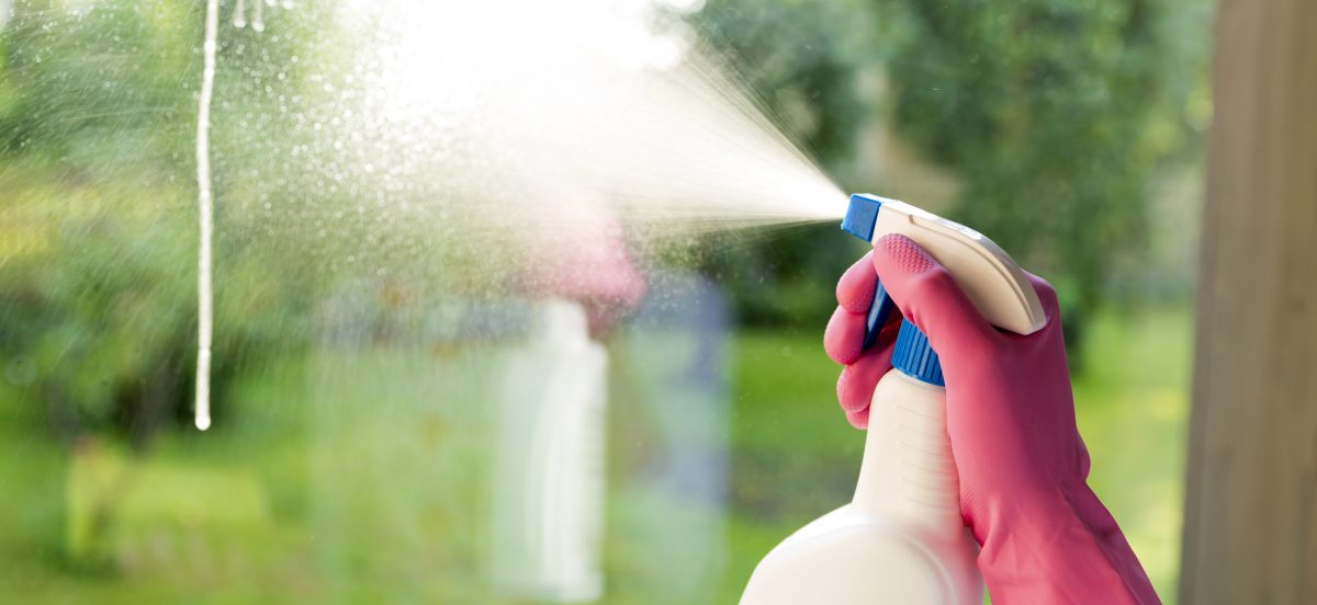 A hand wearing a pink glove sprays cleaning solution from a spray bottle onto a glass window, showing good Product Care outside.