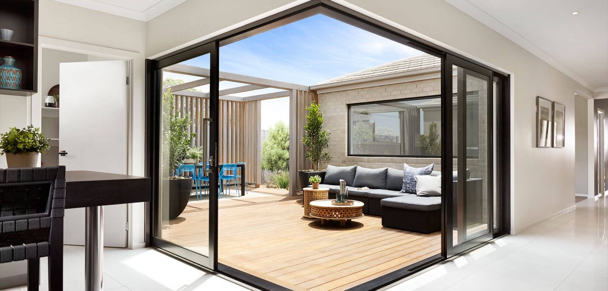 Modern indoor–outdoor living area with glass sliding doors opening to a timber decking, outdoor seating, pot plants, and a dining set under a pergola.