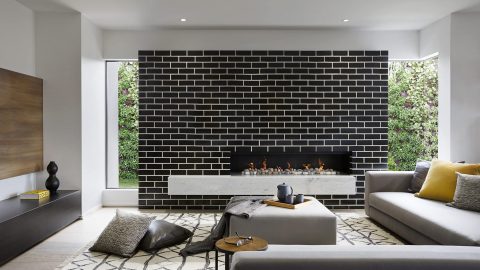Modern living room with a black brick fireplace, grey sofas, geometric rug, low table with a teapot, and floor cushions; large windows and doors look out to lush Aussie greenery.