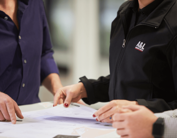 Three people look over documents on a table. One wears a black A&L Services jacket, another points at papers. Faces unseen.