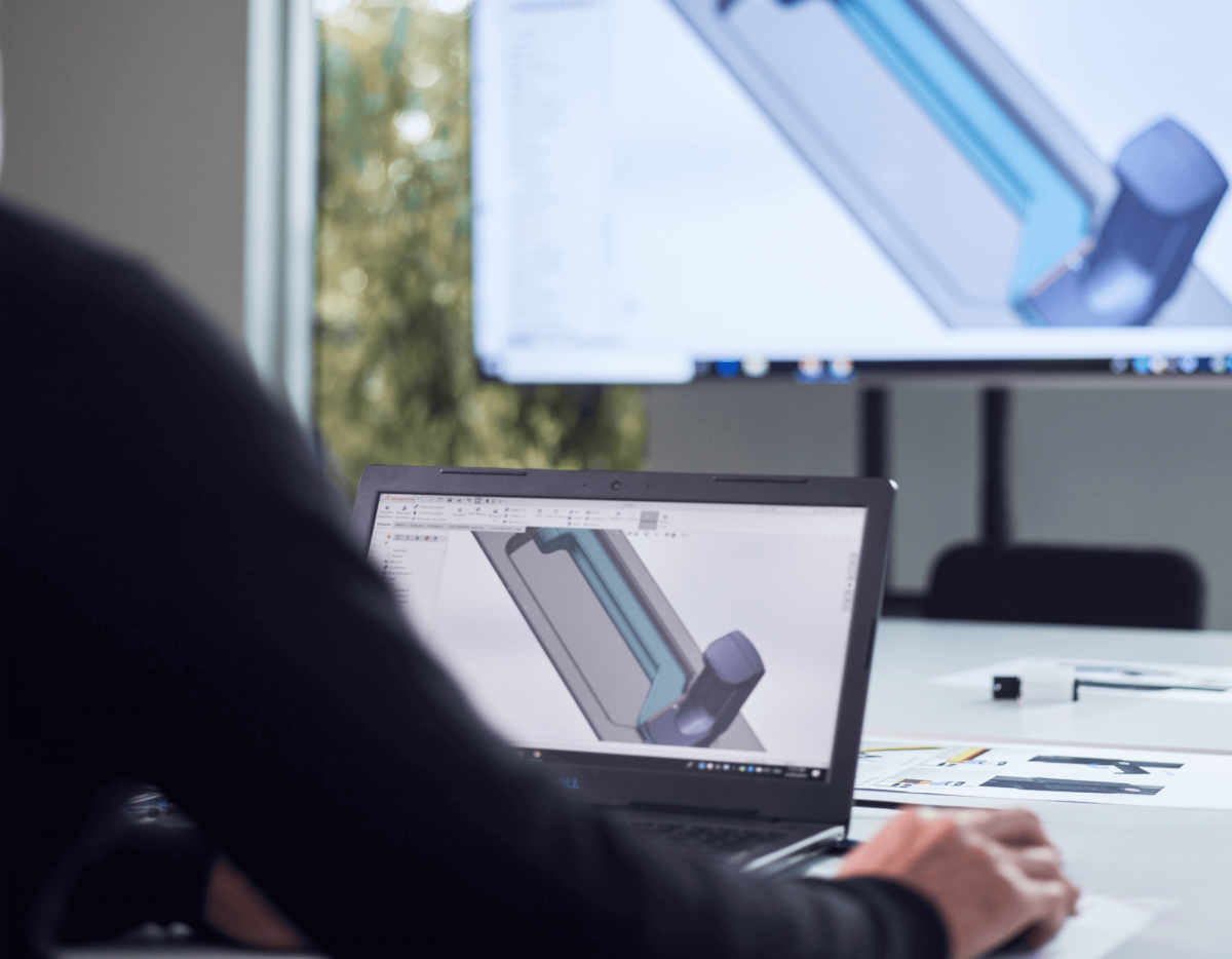 Person working on CAD design on a laptop, same 3D model on a big monitor in a modern office with A&L window visible.