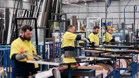 Four Australian workers in yellow safety vests at workstations operate machinery and handle materials inside a large industrial A&L factory.