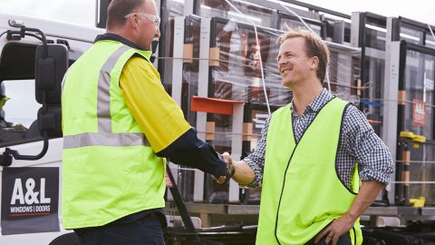 Two men in safety vests shake hands in front of a lorry loaded with A&L window and door frames.