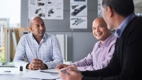 Three men sit at a table in an office with documents; one man smiles. A&L window and door diagrams are on the wall behind them.