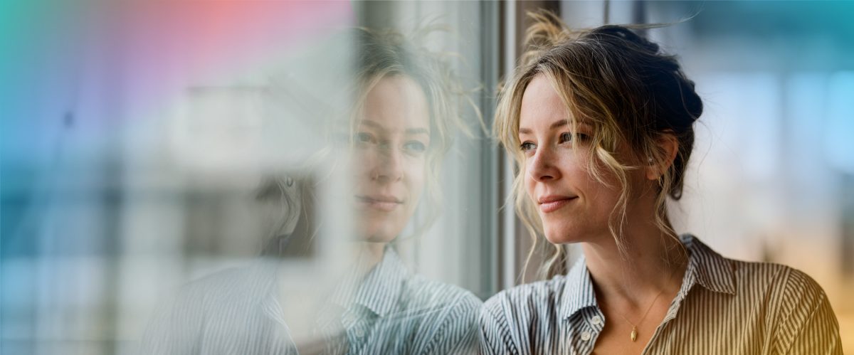 A woman with blonde hair in a striped shirt looks out an A&L window, her reflection blending softly in the glass, framed by A&L doors.
