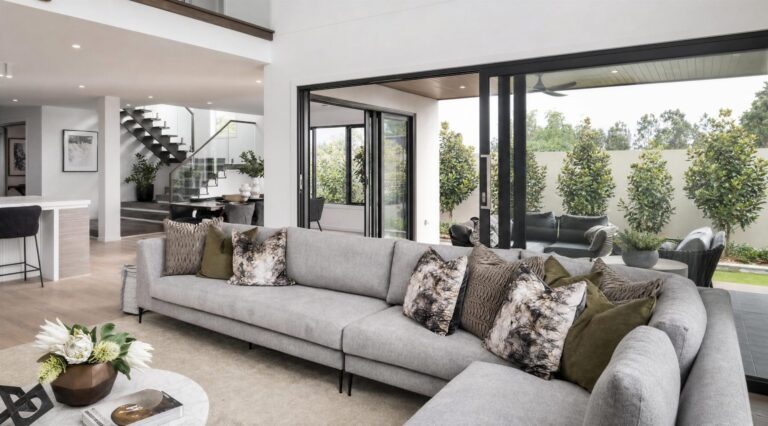 Modern living room with a large grey corner sofa, decorative cushions, and a coffee table with flowers. A&L sliding glass doors open to a patio with outdoor seating and lush greenery.